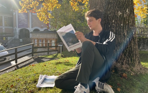 Man sieht einen jungen Mann mit Adidas-Jacke an einem Baum gelehnt sitzen. Es scheint die Sonne. Er liest die Kongresszeitung "Der Moment" am Momentum Kongress 2022