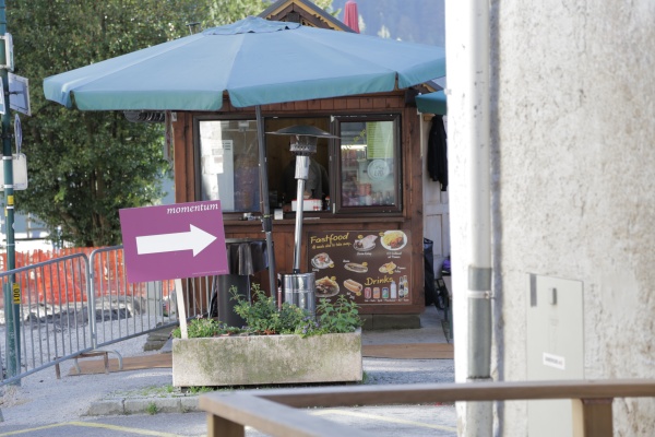 Foto zeigt ein Momentum Schild, das Richtung Kongresszentrum zeigt. Im Hintergrund ist der allseits beliebte Imbissstand, der Kebab, Grillhuhn und Bosna verkauft, zu sehen.
